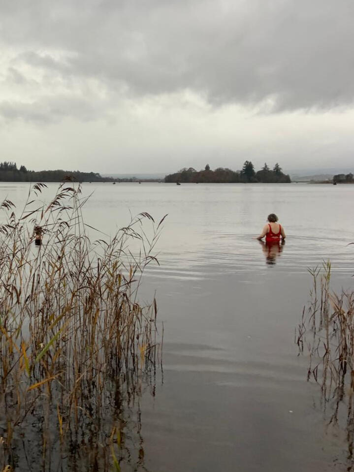 Lisa in a lake in winter A photo of Lisa, a white woman, wearing a red swimsuit , waist high in a lake in autumn. There are pine trees in the distance.